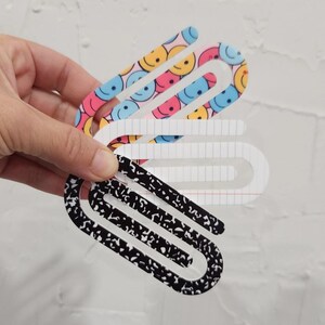 May include: Three colorful paper clips. One is black with white speckles, one is white with a grid pattern, and one is pink with blue and yellow smiley faces.