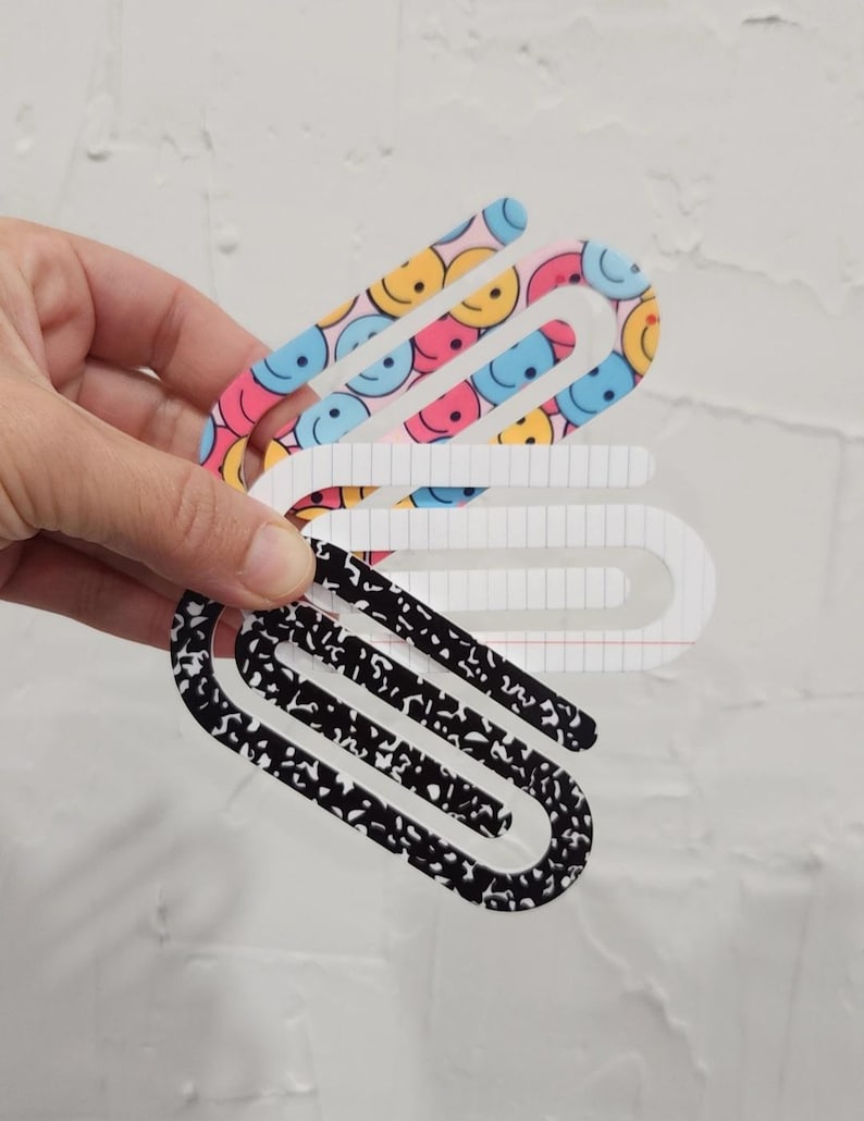 May include: Three colorful paper clips. One is black with white speckles, one is white with a grid pattern, and one is pink with blue and yellow smiley faces.