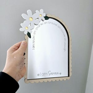 May include: A decorative mirror with a scalloped edge and a floral design. The mirror has the text "I am kind, smart, brave, loved, enough" and "I am Sarina". The flowers are white with gold centres and green leaves.