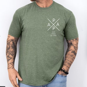 Puede incluir: Una camiseta verde con un dise&ntilde;o gr&aacute;fico blanco. El gr&aacute;fico presenta una cruz con las letras "PAPA" y el texto "Jack Maya Est. 2020".