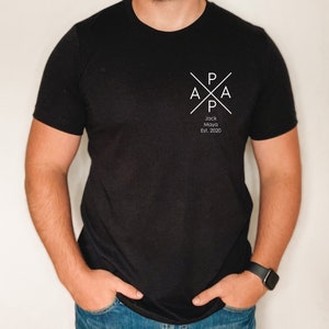 Puede incluir: Camiseta negra con un dise&ntilde;o gr&aacute;fico blanco que presenta las letras "PAPA" en forma de cruz. El texto "Jack Maya Est. 2020" est&aacute; debajo del gr&aacute;fico.