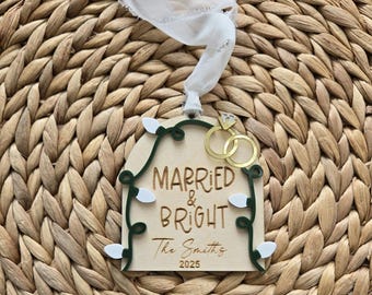 May include: Wooden wedding ornament with the words "Married & Bright" and "The Smiths 2025". The ornament features a gold ring design and is decorated with green and white accents. A white ribbon is attached for hanging.