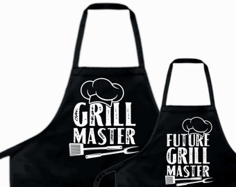 May include: Two black aprons with white text and graphics. The larger apron says "Grill Master" with a chef's hat and grilling tools. The smaller apron says "Future Grill Master" with a chef's hat and grilling tools.