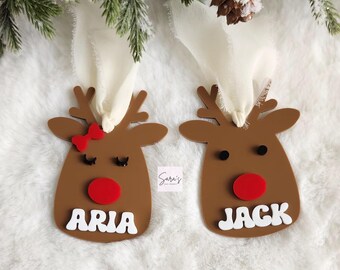 May include: Two brown reindeer ornaments with white names, "Aria" and "Jack", and red noses. Each ornament has a white ribbon attached.