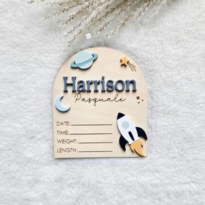 May include: An arched wooden birth announcement sign featuring the name Harrison Pasquale in blue and script lettering. The design includes a planet, a crescent moon, a shooting star, and a rocket ship, with blank lines for date, time, weight, and length.