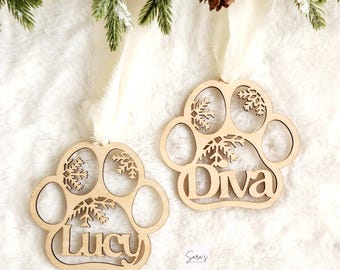 May include: Two wooden paw print ornaments with snowflakes and the names "Lucy" and "Diva" cut out. The ornaments are hanging from white ribbon on a white fuzzy surface.