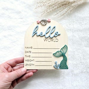 May include: Wooden baby announcement sign with the words "hello WORLD" in blue script. The sign features a mermaid tail, seashell, and stars. Includes spaces to write baby's name, date, time, weight, and length.