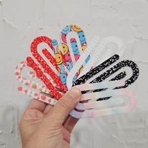 May include: A set of seven colorful paper clips with different patterns, including smiley faces, strawberries, and notebook paper. The paper clips are arranged in a fan shape.