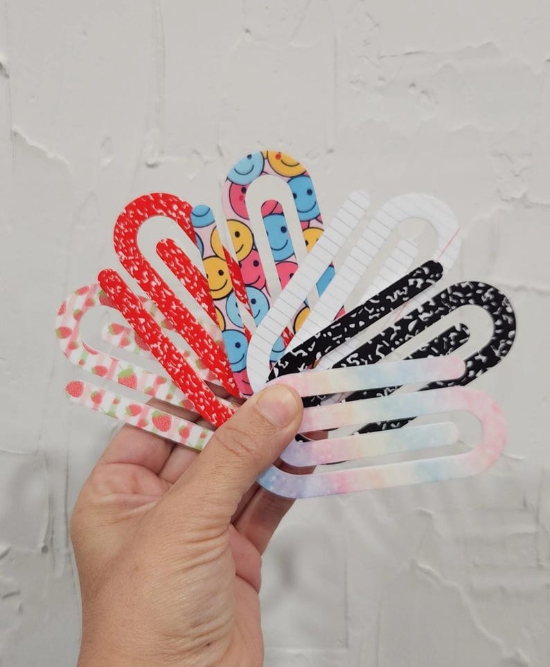 May include: A set of seven colorful paper clips with different patterns, including smiley faces, strawberries, and notebook paper. The paper clips are arranged in a fan shape.