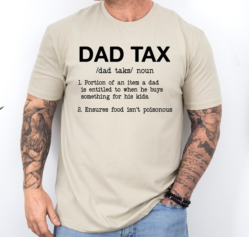 May include: A beige t-shirt with the text "DAD TAX /dad taks/ noun 1. Portion of an item a dad is entitled to when he buys something for his kids. 2. Ensures food isn't poisonous"