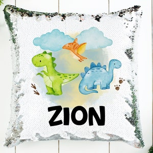 Peut inclure: Un coussin en sequins blanc et argent avec une illustration colorée de trois dinosaures et de nuages. Le texte "ZION" est imprimé en noir sous les dinosaures.