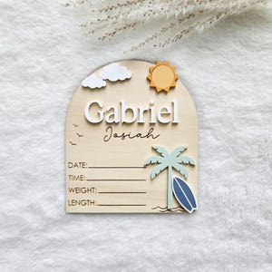 May include: Wooden birth announcement sign with the name "Gabriel Josiah". The sign features a sun, clouds, palm tree, and surfboard design. Includes lines for date, time, weight, and length. The name is in white and black script.