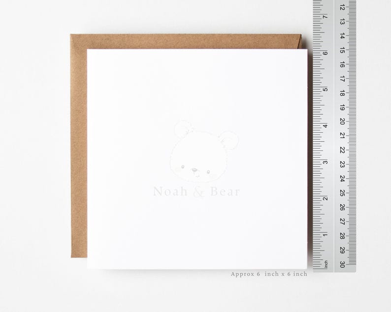 Puede incluir: Una tarjeta de felicitaci&oacute;n cuadrada de color blanco con un dibujo de l&iacute;neas gris claro de una cara de oso y el texto Noah & Bear. Un sobre marr&oacute;n se coloca detr&aacute;s de la tarjeta. Una regla met&aacute;lica muestra que la tarjeta mide unos 15 cm por 15 cm.