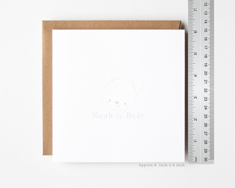 May include: A square white greeting card with a light gray line drawing of a bear face and the text Noah & Bear. A metal ruler is placed vertically next to the card, showing dimensions of approximately 6 inches by 6 inches.
