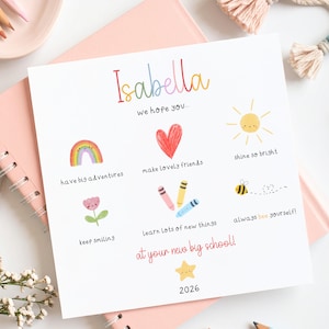 May include: A square white card featuring the name Isabella in colourful letters, with illustrations of a rainbow, heart, sun, flower, crayons, and bee. Text includes: we hope you have big adventures, make lovely friends, shine so bright, keep smiling, learn lots of new things, always bee yourself, at your new big school, and 2026.