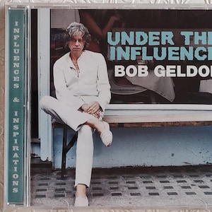 May include: CD case for Bob Geldof's album "Under the Influence." The cover features a man in white clothing seated on a bench. The album title is in turquoise block letters, with the artist's name below.