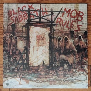 May include: Album cover art for Black Sabbath's "Mob Rules" featuring a brick wall with the band's name and album title in red. Figures in hooded cloaks stand before a tattered banner. Artwork by Greg Hildebrandt.
