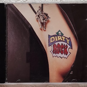 May include: CD case featuring a close-up of a person's arm with a "Dirty Rock" tattoo in blue, red, and white. A silver bolo tie with a detailed pendant hangs near the arm. The person is wearing a black leather jacket.
