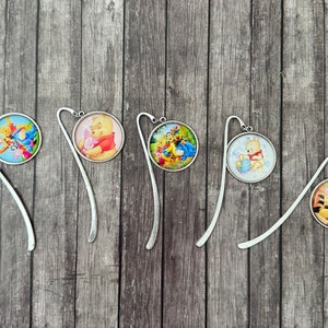 Puede incluir: Cinco marcapáginas de metal plateado con diferentes personajes de Winnie the Pooh en el encanto redondo de vidrio. Los encantos son coloridos y presentan a Winnie the Pooh, Piglet, Tigger y Eeyore.
