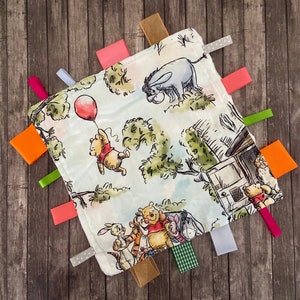 May include: A white baby blanket with colourful fabric tags. The blanket features a Winnie the Pooh and friends theme with illustrations of Winnie the Pooh, Piglet, Eeyore, Tigger, and a donkey.