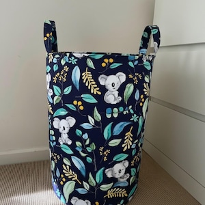 Può includere: Un cesto portabiancheria cilindrico con sfondo blu navy, con un motivo di koala, foglie verdi e bacche gialle. Il cesto ha due manici in tessuto ed è progettato per la conservazione.