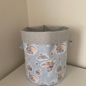 May include: A blue and grey fabric storage bin with a pattern of Dumbo the elephant flying. The bin has a grey polka dot lining.