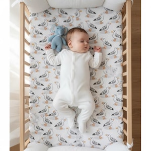 May include: A baby in a white onesie lies in a wooden crib on a fitted sheet patterned with gray seagulls. A blue stuffed elephant toy rests near the baby. The crib has a white bumper and is set against a neutral background.
