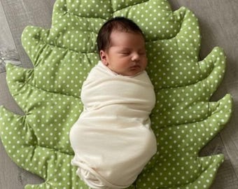 May include: A newborn infant with dark hair is wrapped in a plain off-white swaddle blanket. The infant is lying on a padded, leaf-shaped mat that is light green with a repeating pattern of small white polka dots and stitched vein details.