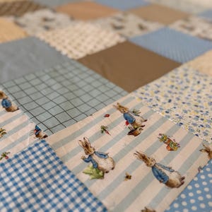 以下が含まれることがあります： 青、ベージュ、白の色合いの様々な布の正方形を組み合わせたパッチワークキルト。ギンガムチェック、水玉模様、格子柄、花柄、そして青いジャケットを着たピーターラビットのイラストなど、多様なパターンがデザインされています。