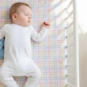 Op de afbeelding: Een slapende baby in een witte onesie ligt op een pastel geruit beddengoed naast een witte wieg. Een klein blauw knuffeldier is zichtbaar bij de baby. Het beddengoed heeft een patroon van lichtroze, gele, blauwe en groene vierkanten.