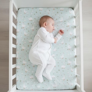 May include: A sleeping baby in a white onesie lies in a white crib on a light blue fitted sheet. The sheet is patterned with white geese and green grass. The crib is white.