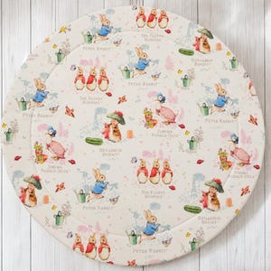 May include: A round, cream-coloured fabric mat featuring a repeating pattern of Beatrix Potter characters including Peter Rabbit, Benjamin Bunny, The Flopsy Bunnies, and Jemima Puddle-Duck, interspersed with watering cans, cucumbers, and flowers.