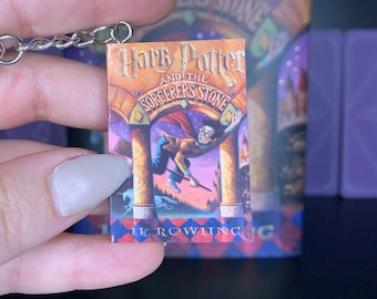 May include: Miniature keychain featuring the cover art of "Harry Potter and the Sorcerer's Stone" by J.K. Rowling. The book cover shows Harry Potter on a broomstick. The keychain is held in front of a larger book with the same title.