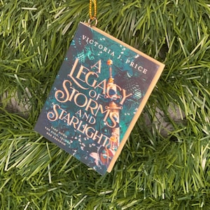 May include: A book ornament featuring a green and blue cover with the title "A Legacy of Storms and Starlight" by Victoria J. Price. The ornament is hanging from a gold string.