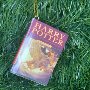May include: A red and white book ornament with a gold string hanging from the top. The book cover features an illustration of a creature with wings and a person riding on its back. The title of the book is "Harry Potter and the Prisoner of Azkaban" by J.K. Rowling.