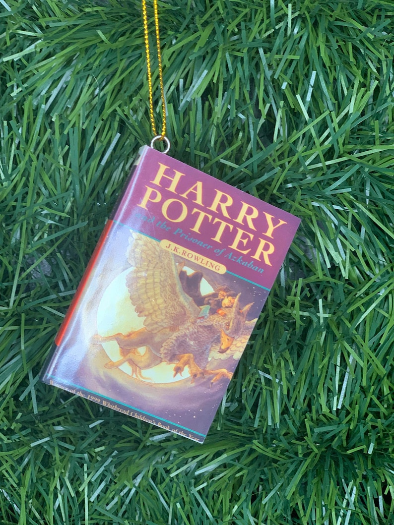 May include: A red and white book ornament with a gold string hanging from the top. The book cover features an illustration of a creature with wings and a person riding on its back. The title of the book is "Harry Potter and the Prisoner of Azkaban" by J.K. Rowling.