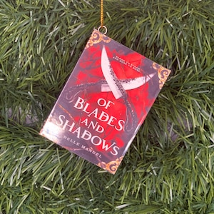 May include: A red book with a gold design on the cover. The title is "Of Blades and Shadows" by Belle Manuel. The book is hanging from a gold string.