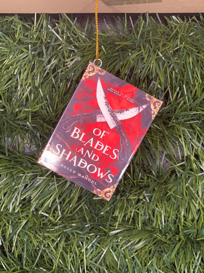 May include: A red book with a gold design on the cover. The title is "Of Blades and Shadows" by Belle Manuel. The book is hanging from a gold string.