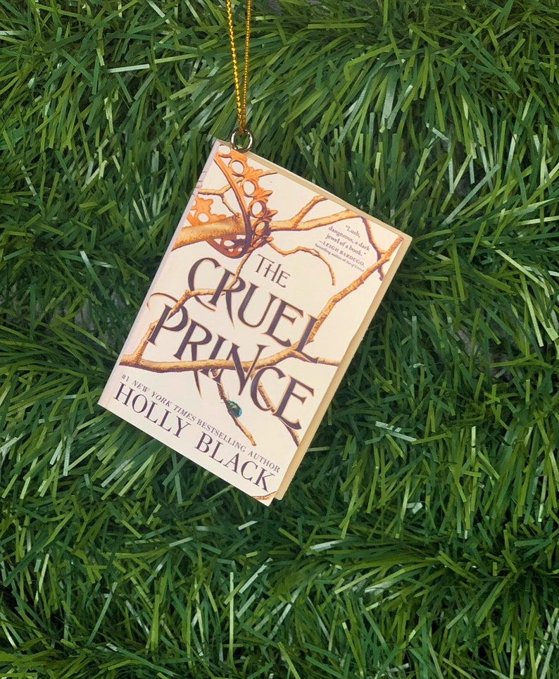 May include: A white and gold Christmas ornament shaped like a book with the title "The Cruel Prince" by Holly Black. The ornament features a stylized illustration of a tree branch with thorns.