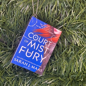 May include: A blue and black book ornament with a silver chain. The book cover features the title "A Court of Mist and Fury" by Sarah J. Maas.