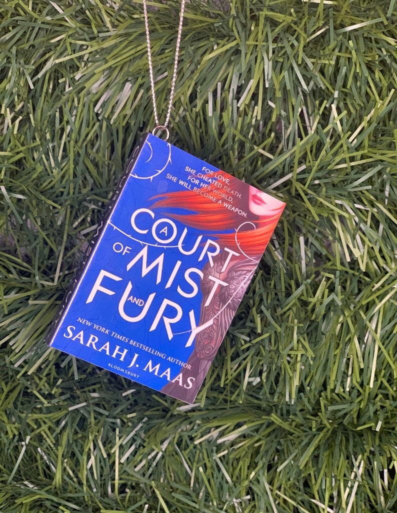 May include: A blue and black book ornament with a silver chain. The book cover features the title "A Court of Mist and Fury" by Sarah J. Maas.