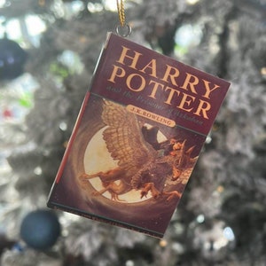 May include: A red and gold Harry Potter book ornament featuring the title "Harry Potter and the Prisoner of Azkaban" by J.K. Rowling. The ornament depicts a winged creature with a rider.