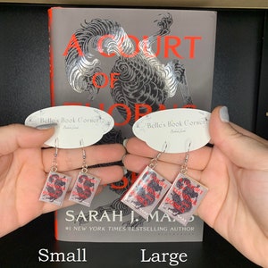 May include: A pair of silver earrings with a book charm. The book cover features a black and white illustration of a dragon with the text "A Court of Thorns and Roses" and "Sarah J. Maas" in red. The earrings are shown in two sizes: small and large.