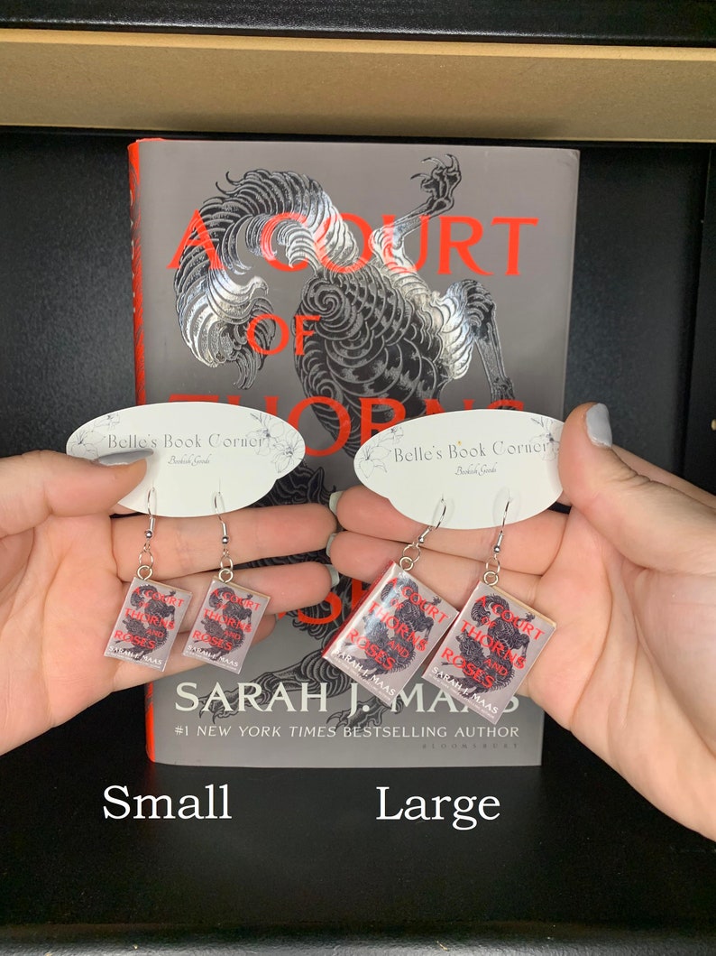 May include: A pair of silver earrings with a book charm. The book cover features a black and white illustration of a dragon with the text "A Court of Thorns and Roses" and "Sarah J. Maas" in red. The earrings are shown in two sizes: small and large.