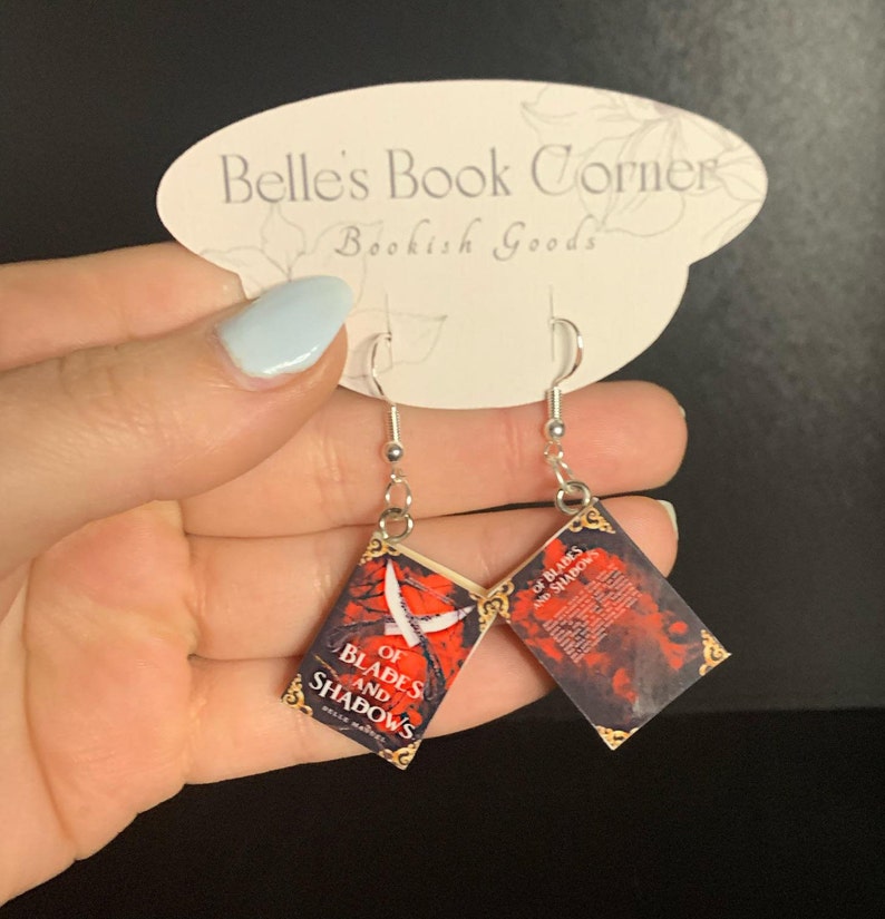 May include: A pair of silver earrings with miniature book charms featuring the cover of the book "Of Blades and Shadows" by  Cassandra Clare. The book covers are red, black, and gold.