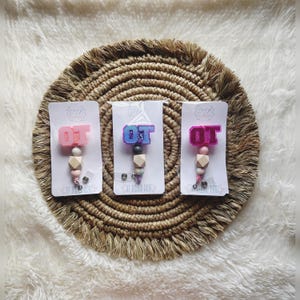 Puede incluir: Tres abalorios de letras 'OT' de colores con cuentas de madera y un carrete de insignia retráctil. Los abalorios son rosa, azul y morado con detalles de purpurina. Los abalorios están sobre un fondo blanco con el texto 'OT Hugs' en las tarjetas.