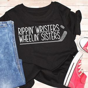 Puede incluir: Camiseta negra con texto blanco que dice "Rippin' Wristers Wheelin' Sisters" y un gráfico de un palo de hockey.