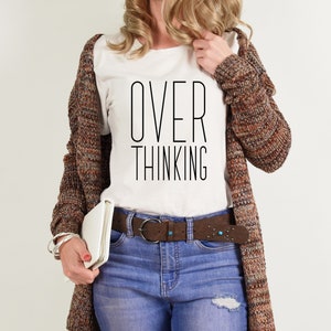 May include: White t-shirt with the text "OVER THINKING" in black lettering. The shirt is worn by a person with a brown cardigan and blue jeans.