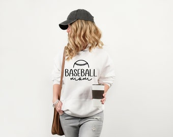 以下が含まれることがあります： 黒い野球のグラフィックと黒い筆記体で「Baseball mom」という文字が書かれた白いフーディー。モデルは黒い野球帽とグレーのジーンズを着用しています。