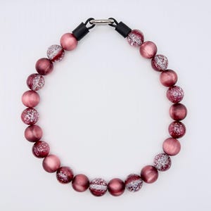 May include: A necklace featuring large, round beads in shades of pink and burgundy. Some beads have a glittery, silver-colored design. The necklace is connected with a silver-colored clasp and black connectors.